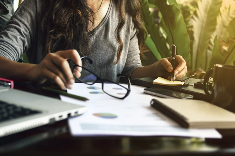 A woman (face unseen) sitting a desk with a notepad and glasses in hand.