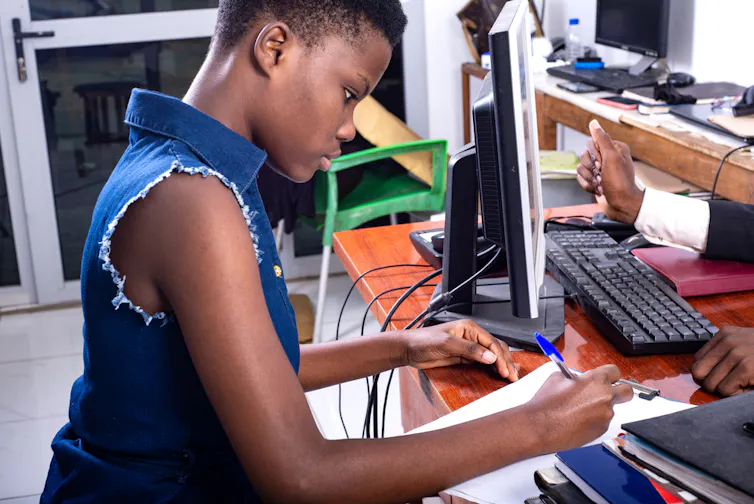 A young girl sitting a desk with a notebook and pen deep in concentration.