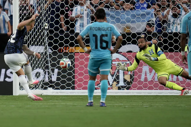 A soccer player stands facing a net while a goalie dives for a ball.