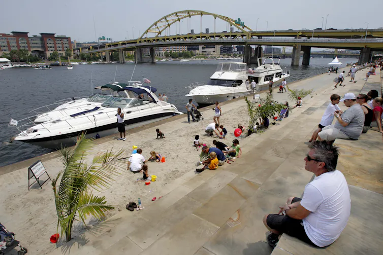 A group of people sitting along the bank of a river full of boats.