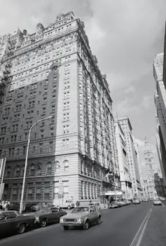 Black-and-white photo of large ornate building alongside two-way street with 1970s-era cars