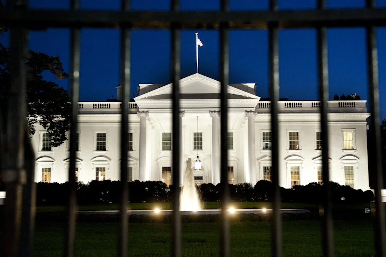 a large white building lit up at night seen through an iron fence