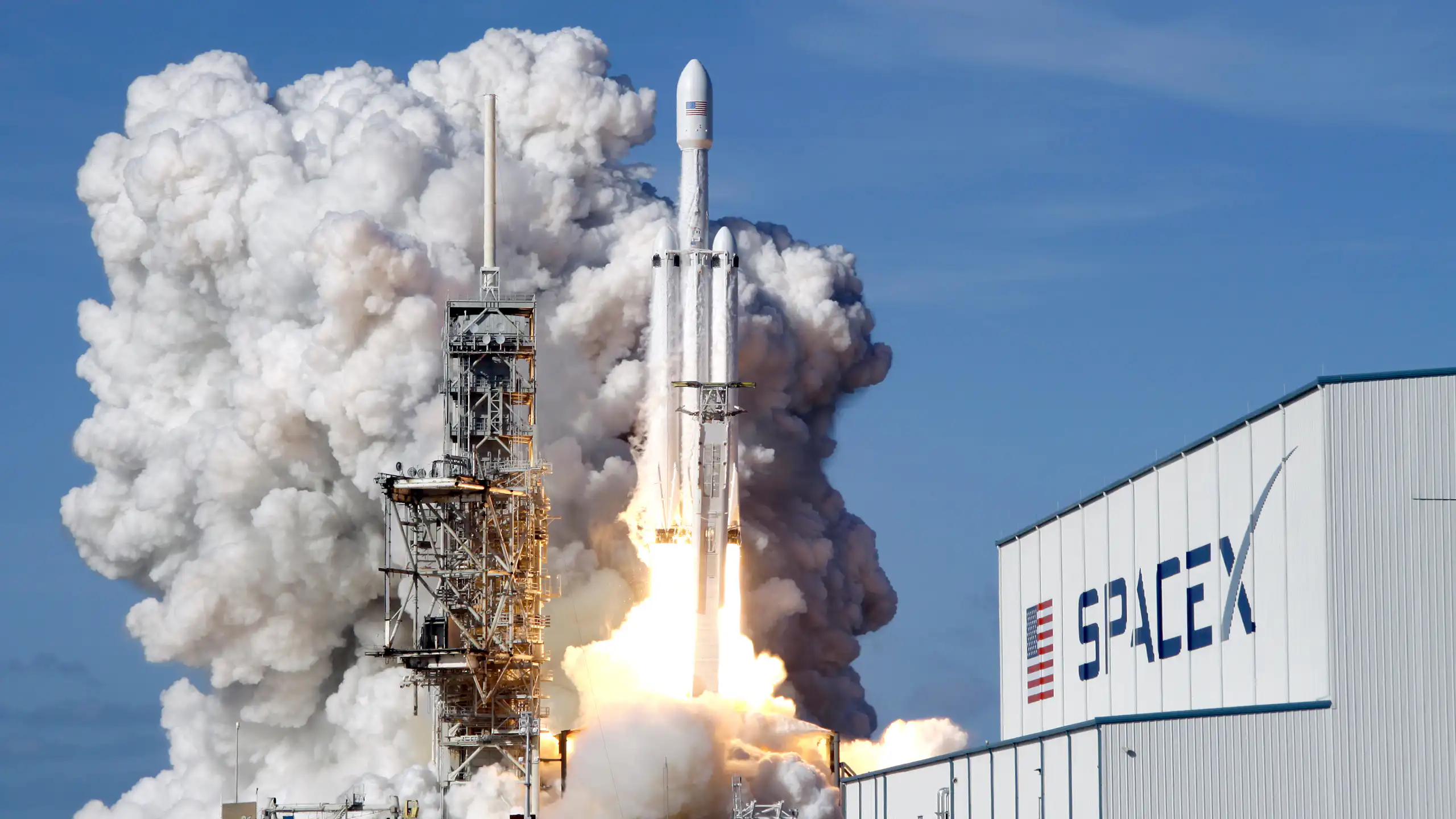 A Falcon 9 SpaceX heavy rocket can be seen lifting off amid a engine fire at the bottom and billowing gray smoke next to a building that says spacex