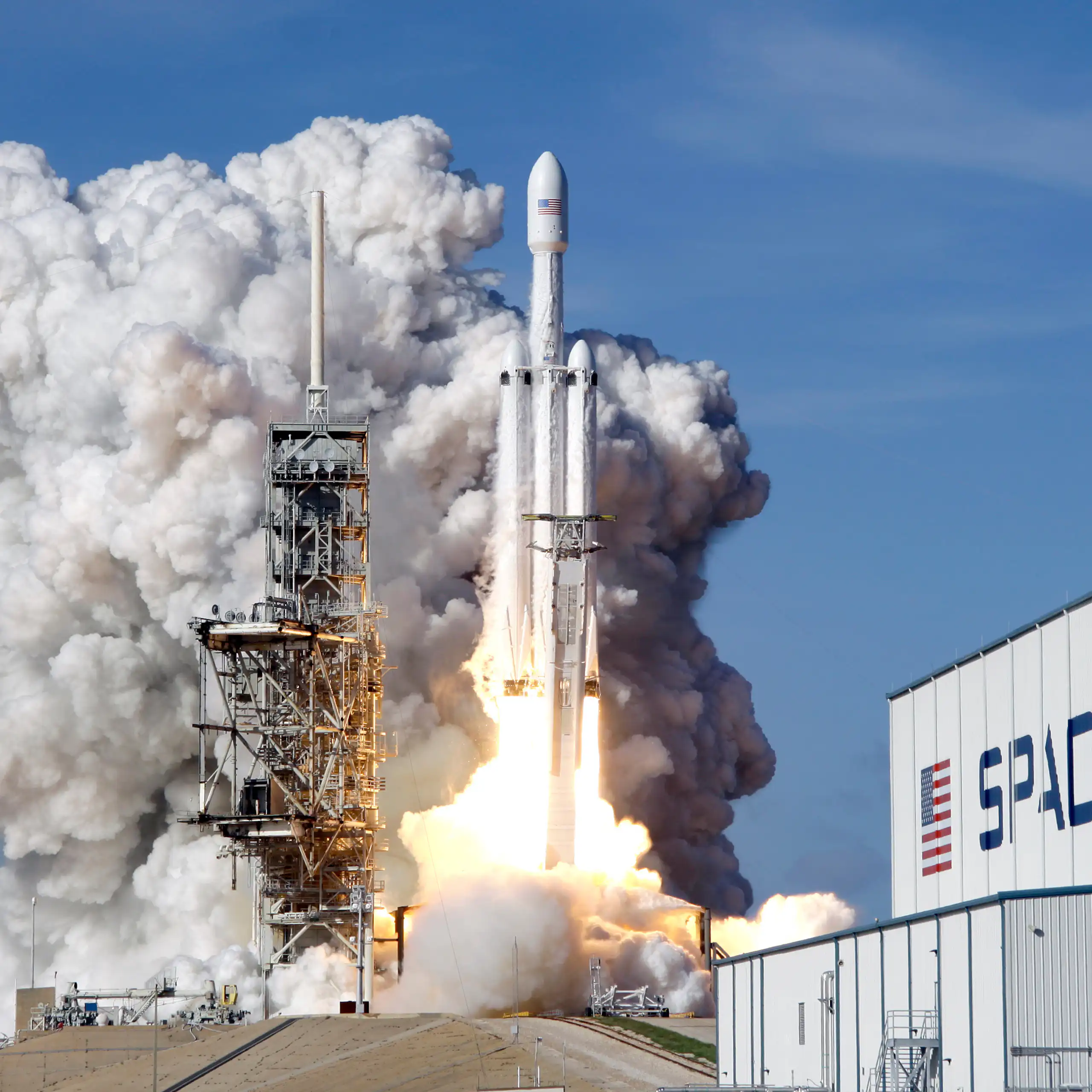 A Falcon 9 SpaceX heavy rocket can be seen lifting off amid a engine fire at the bottom and billowing gray smoke next to a building that says spacex