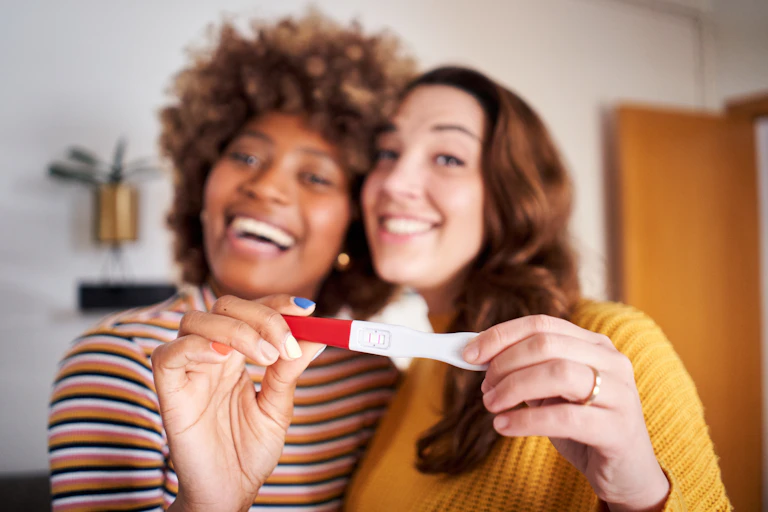Two smiling women standing closely together hold up a positive pregnancy test to the camera
