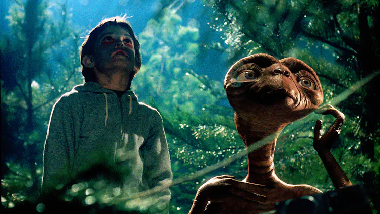 Un niño y un extraterrestre en medio del bosque miran hacia el cielo.
