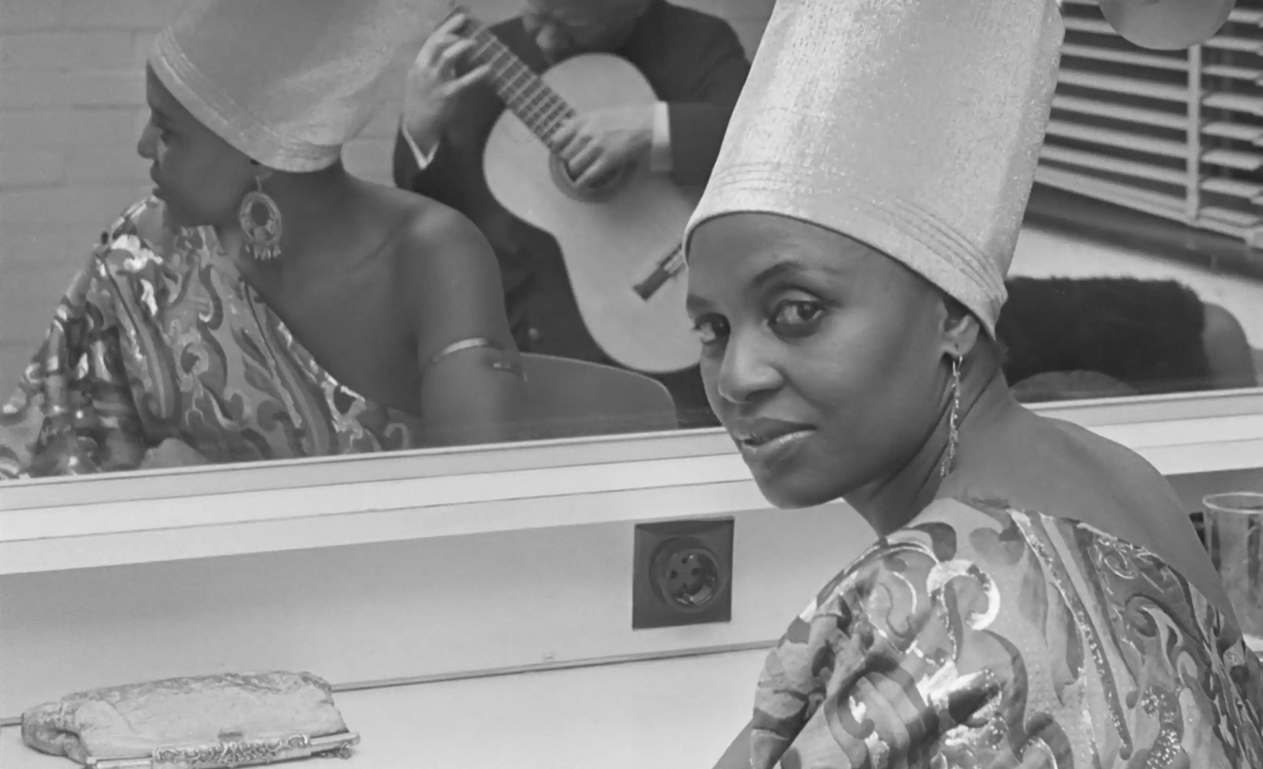 A young African woman sits in front of a mirror in a glamorous outfit with elongated headgear. She looks frankly to camera. In the mirror is a reflection of a man playing a guitar.