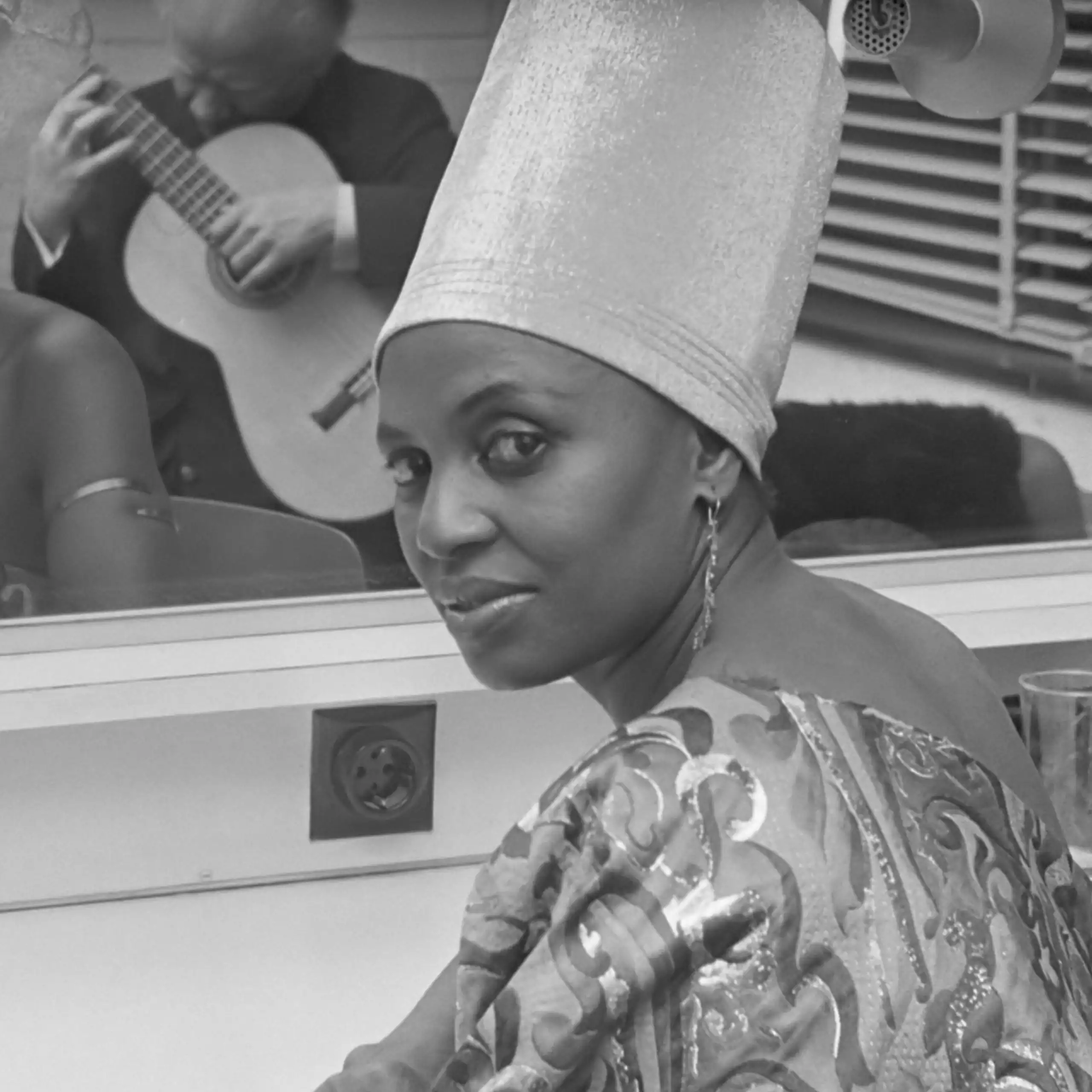 A young African woman sits in front of a mirror in a glamorous outfit with elongated headgear. She looks frankly to camera. In the mirror is a reflection of a man playing a guitar.