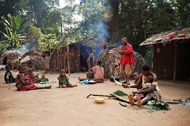 Children sit on the ground in various poses. Some cook and help with food prep. A man in a red shirt stands with his arms folded, watching.