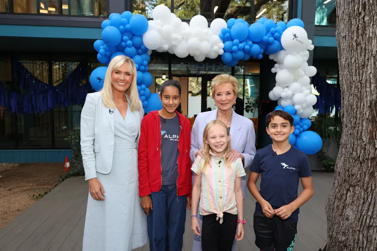 Two women with blonde hair stand together and smile with three children near a blue-and-white balloon arch outside of a building.