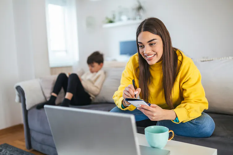 A woman smiling in front of a laptop while a child reads in the background