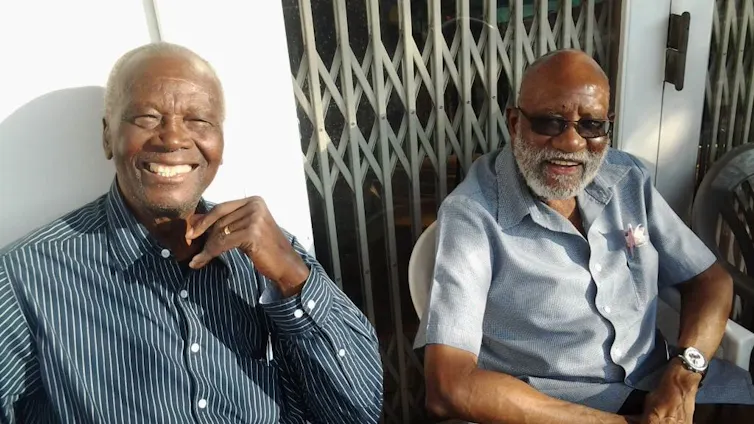 Two elder African men sit in the sun outside a house, both smiling broadly at the camera.