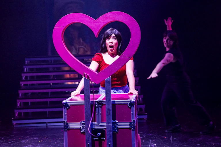 A theatre performer onstage has their head framed inside a pink heart. Another cast member dances in the background.