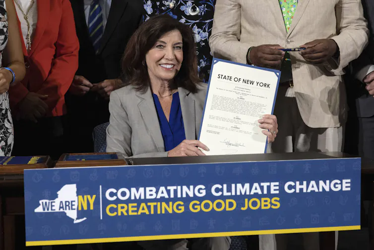 The governor smiles as she holds the signed legislation, sitting in front of a banner reading 'Combating climate change, creating good jobs'.
