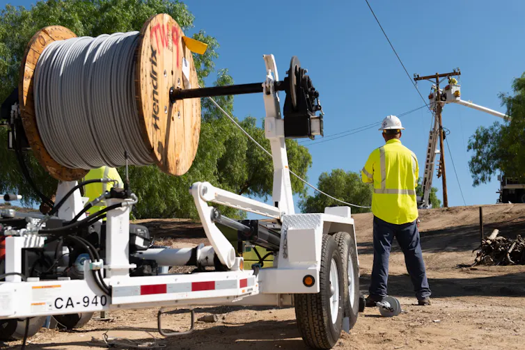 A worker stands by a giant roll of covered conductor line – power lines covered in a plastic.