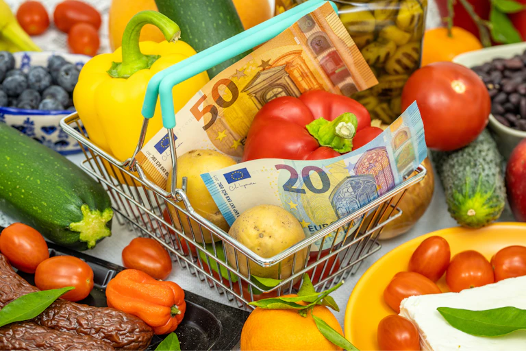 Cesta de la compra con verduras y billetes de euro.