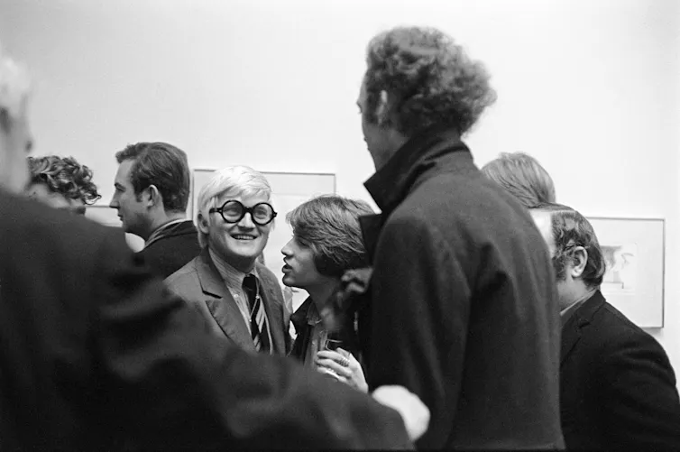 A group of people at an art gallery with David Hockney in the middle, smiling.