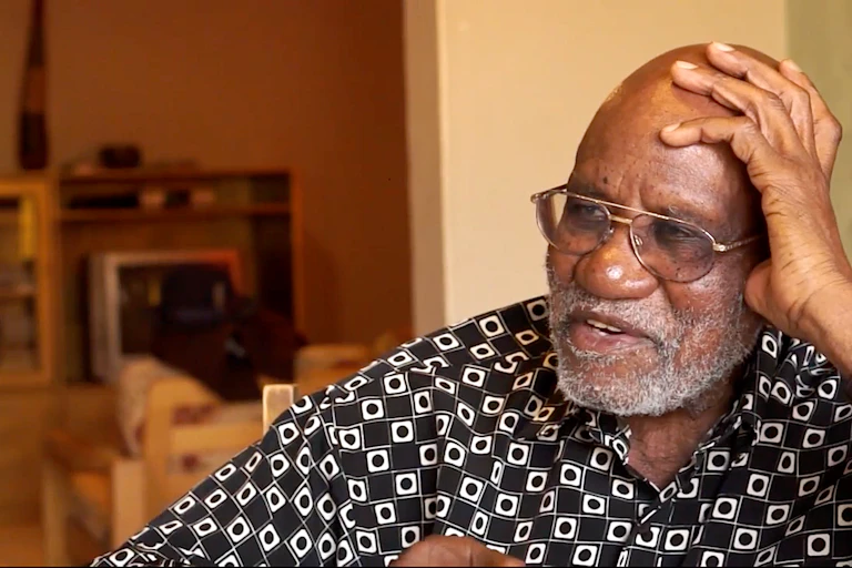 An elder African man sits talking, a hand on his bald head above a white beard and spectacles, dressed in a striking black and white patterned shirt.