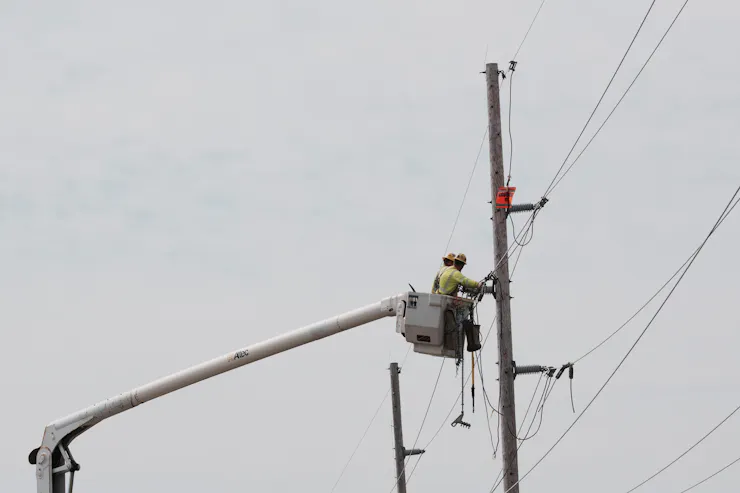 A person wearing a hard hat stands in a raised bucket and works on power lines attached to a large pole.