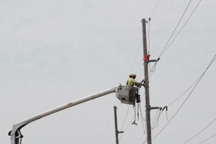 A person wearing a hard hat stands in a raised bucket and works on power lines attached to a large pole.