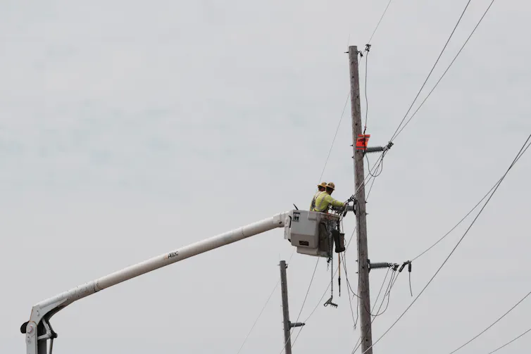A person wearing a hard hat stands in a raised bucket and works on power lines attached to a large pole.