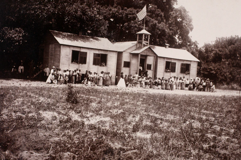 A black and white photo shows a large group of people of color outside of a one-level white building with a flag on top.