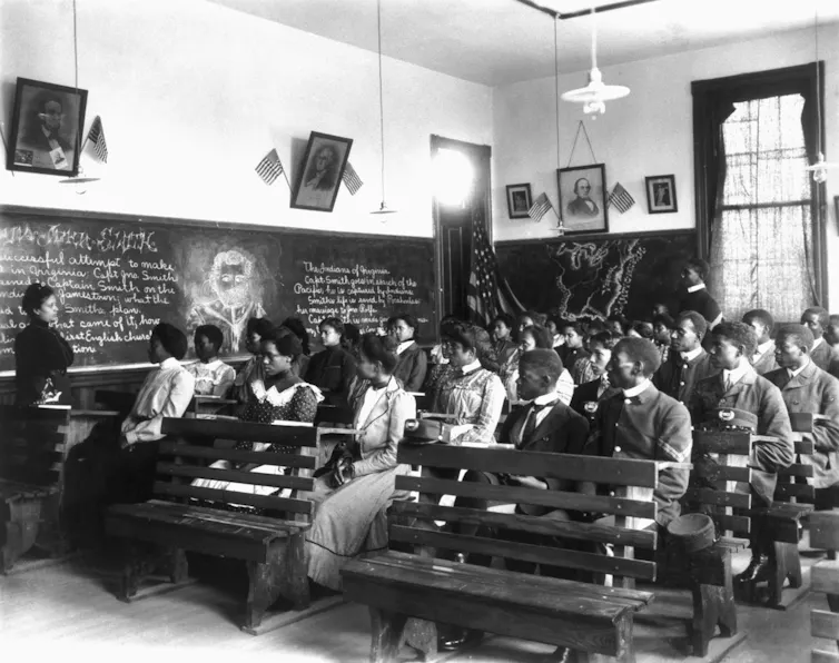 Juneteenth nos recuerda la larga lucha de los afroamericanos por la educación después del fin de la esclavitud 1 La fotografía en blanco y negro muestra varias filas de jóvenes negros vestidos formalmente sentados en filas de bancos con una pizarra cerca.