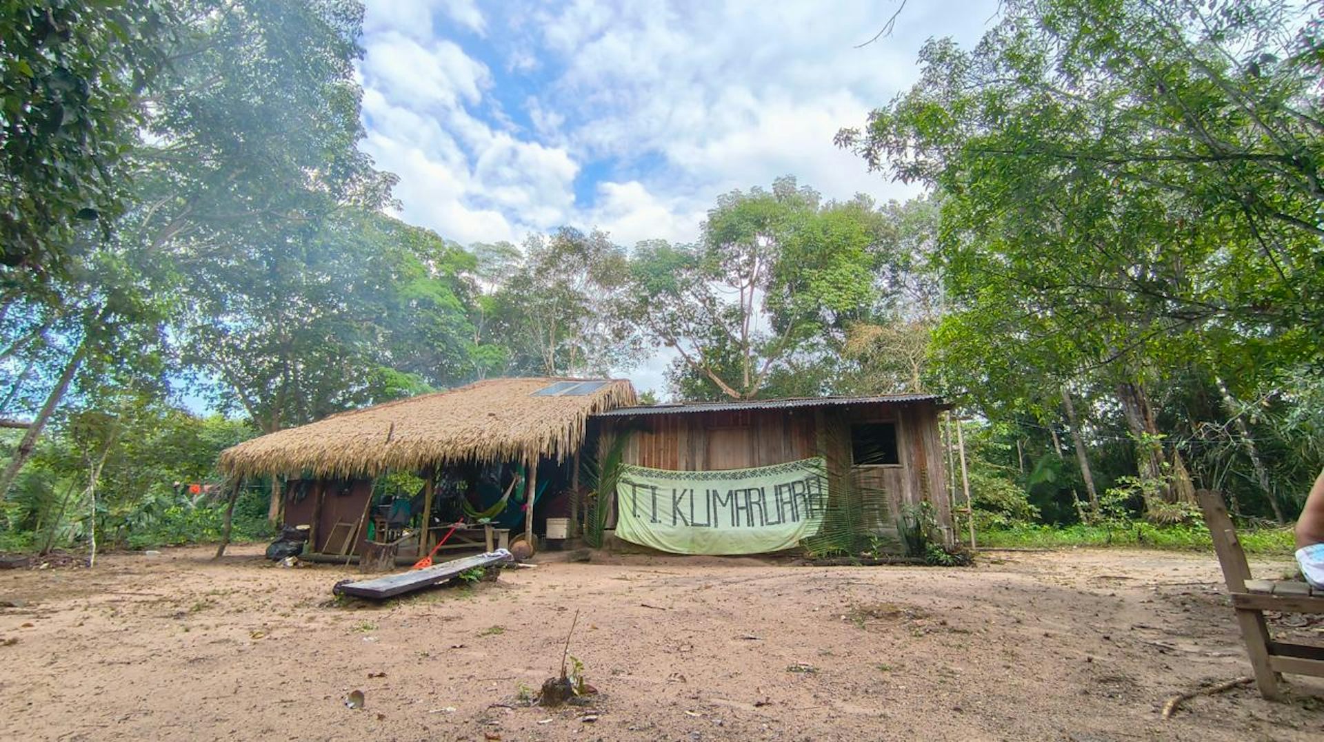 Foto mostra uma casa de madeira e palha no meio da floresta. Na casa, uma faixa escrita T.I. Kumaruara