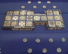 Tablero de juego de madera; la superficie está formada por 20 placas cuadradas de concha con incrustaciones variadas; los bordes están hechos de pequeñas placas y tiras, algunas esculpidas con un ojo y otras, posiblemente, con rosetas; en el reverso hay t