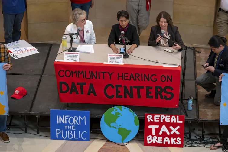 People sit at a table decorated with signs saying 'community hearing on data centers,' 'inform the public' and 'repeal tax breaks.'