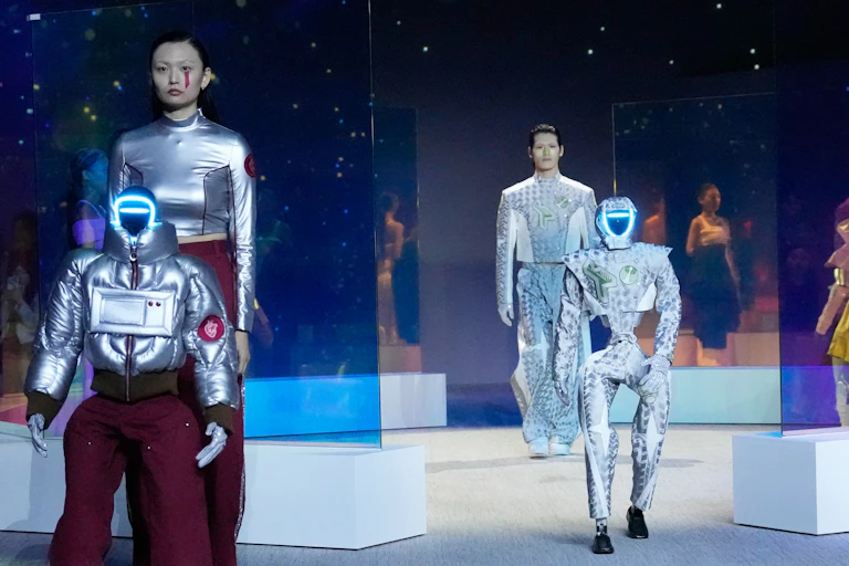 A woman and man dressed in futuristic silver clothing stands expressionless behind two humanoid robots in similar clothing on the catwalk.