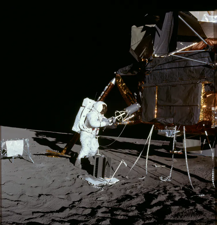 An astronaut in a white spacesuit tending to machines on a grey, dusty moon.