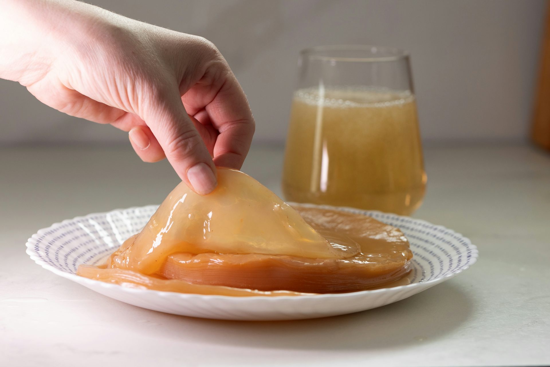 A hand lifts the SCOBY from a plate. A glass of freshly brewed kombucha sits nearby.