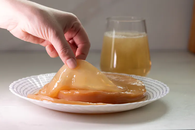 A hand lifts the SCOBY from a plate. A glass of freshly brewed kombucha sits nearby.