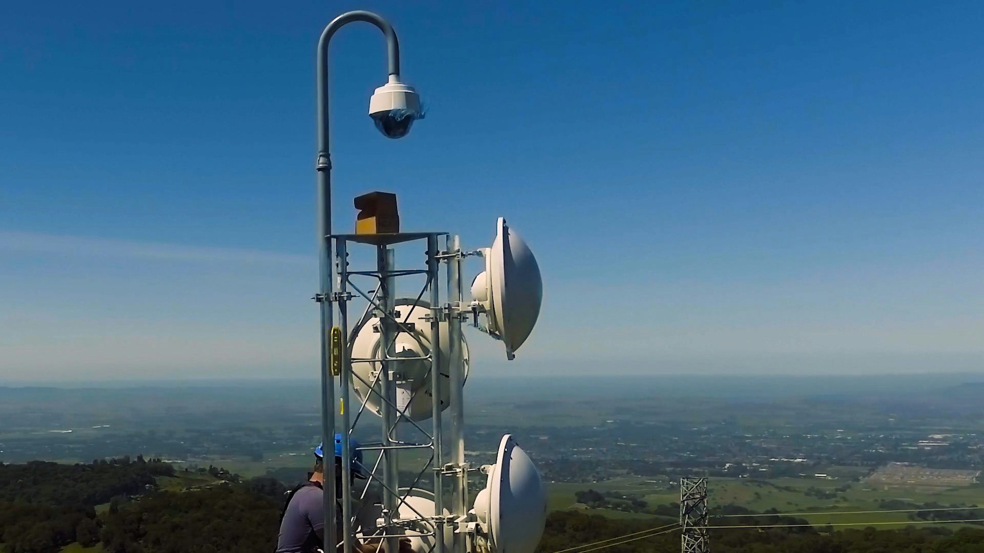 Un trabajador trepa hacia la cámara por un poste con platos de microondas a su alrededor.