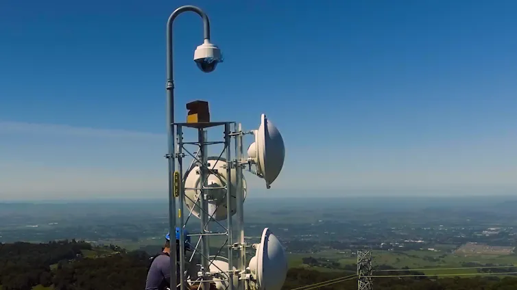 A worker climbs toward a camera on a pole with microwave dishes around it.