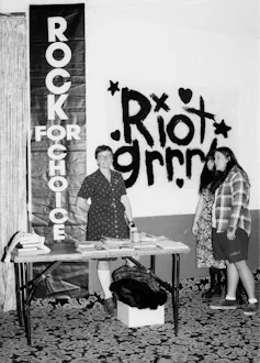 Black-and-white photo of three young women standing near a display table featuring a range of publications.