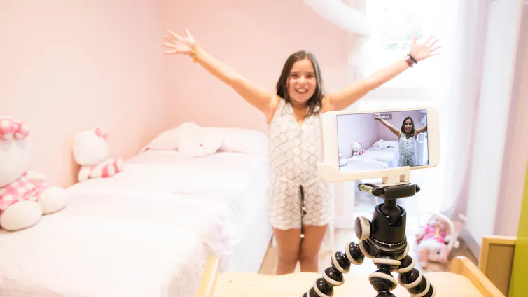 A young girl is seen with her arms up in a pink bedroom. She looks at a smartphone mounted on a table that is filming her.