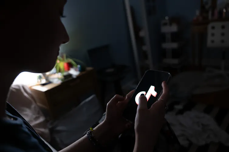 A young person is seen in the shadows looking at a smartphone that has a TikTok image on it.
