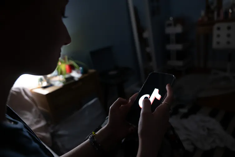 A young person is seen in the shadows looking at a smartphone that has a TikTok image on it.
