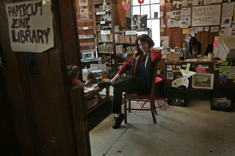 A young woman seated in a chair in a wood paneled room surrounded by shelves with zines and a wall with various images and flyers tacked to it.