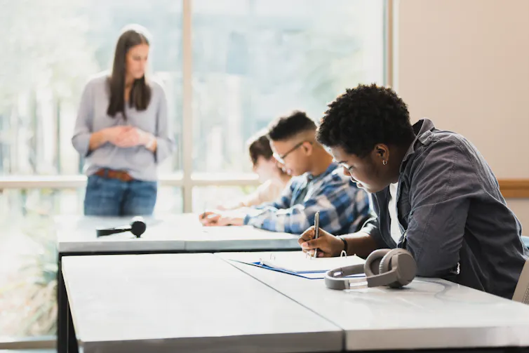 Three students are seen in a row leaning over papers on a table as a woman stands nearby them.