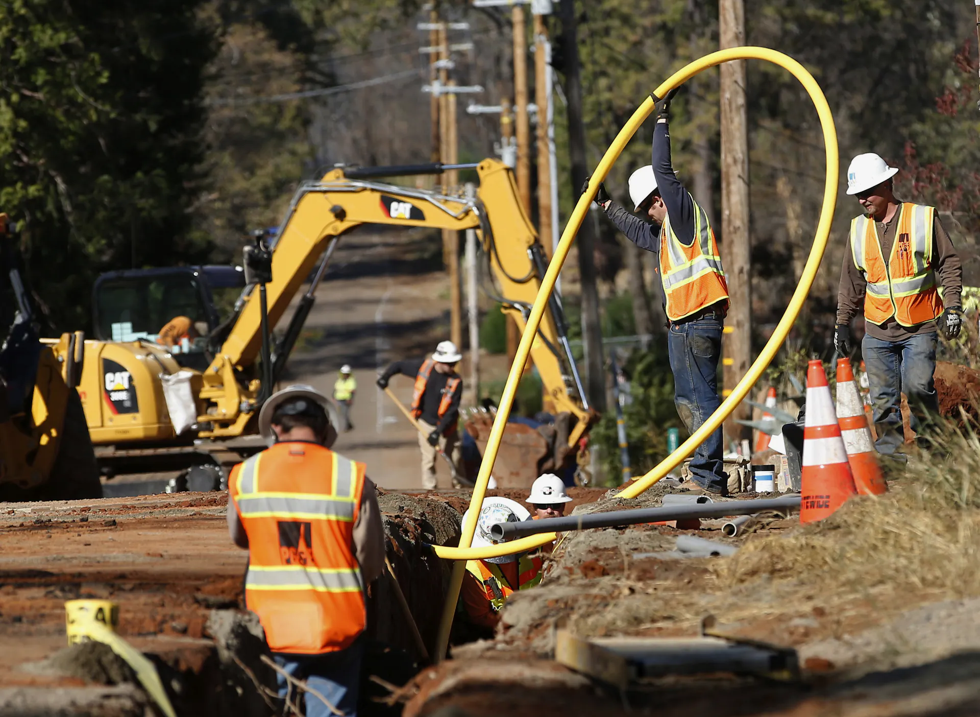 Utility workers in hard hats and reflective vests burying a power line in a trench next to a road.