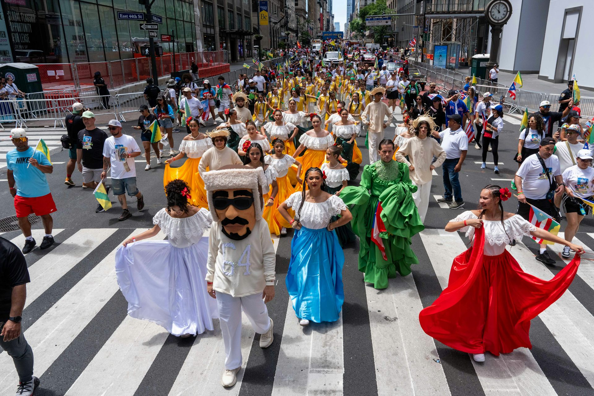Dancers in a parade stream through a street and one is wearing a costume of a man in a suit in sunglasses.