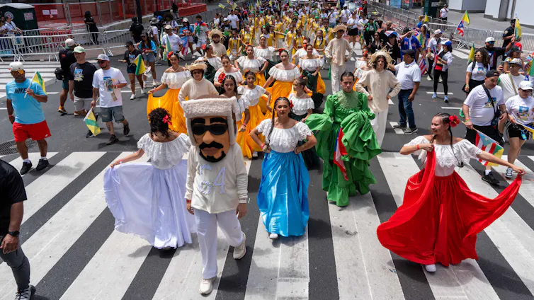 music Dancers in a parade stream through a street and one is wearing a costume of a man in a suit in sunglasses.