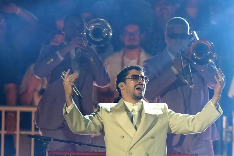 A man in a white suit jacket and dark sunglasses singing with his hands up in front of players with brass instruments.