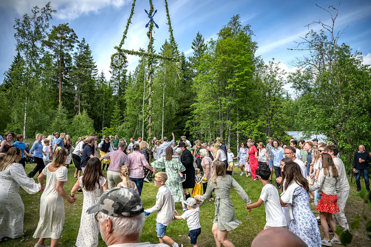 A large group of people holding hands dances in a circle outside around a large pole wound with greenery.