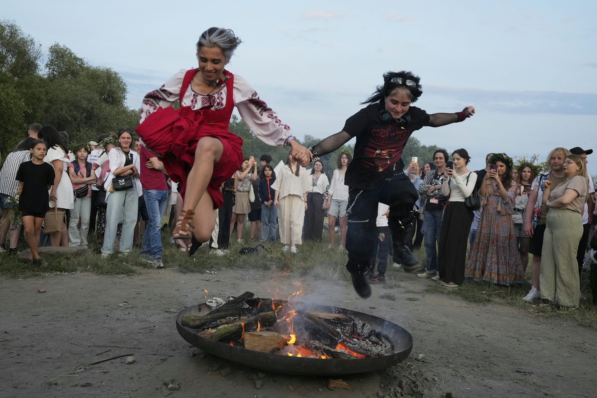Dos mujeres, una de ellas con un vestido rojo tradicional, se toman de la mano mientras saltan sobre una pequeña llama afuera.