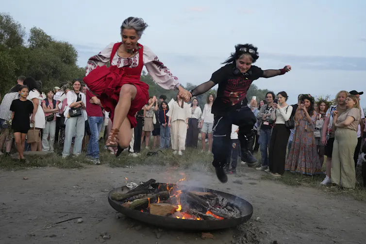 Hogueras, mayos y santo: cómo Europa celebra el día más largo del año 1 Dos mujeres, una de ellas con un vestido rojo tradicional, se toman de la mano mientras saltan sobre una pequeña llama afuera.