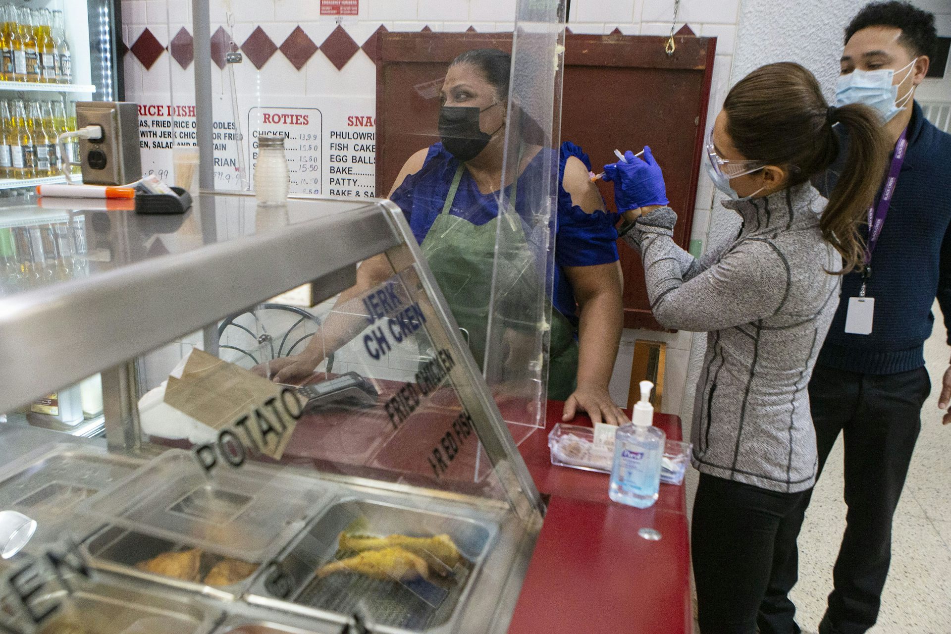 Una mujer con mascarilla le da una inyección a una mujer parada cerca de un mostrador de comida rápida.